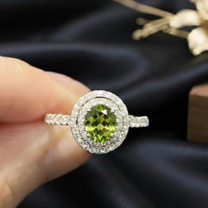 Könnte beinhalten: Ein silberner Ring mit einem oval geschliffenen, hellgrünen Edelstein, umgeben von zwei Ringen aus kleineren, klaren Edelsteinen. Der Ring hat ein mit kleinen, klaren Edelsteinen besetztes Band. Der Ring wird vor dunklem Hintergrund gehalten.