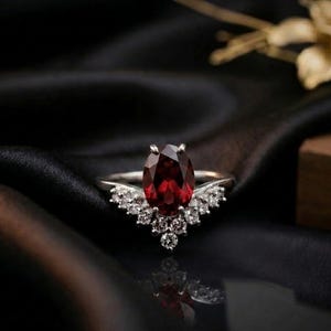 May include: A silver ring with a large, oval-cut, deep red gemstone as the centerpiece. The ring is accented with smaller, clear gemstones in a decorative pattern. The ring is set against a dark background.