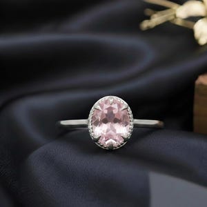 May include: A silver ring with a large, oval-shaped, light pink gemstone. The gemstone is set in a detailed bezel setting. The ring is displayed on a dark, textured fabric.