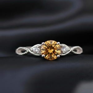 May include: A silver ring with a large, round, golden-yellow gemstone at the center, flanked by two smaller white gemstones. The band features a twisted design. The ring is set against a dark, textured background.