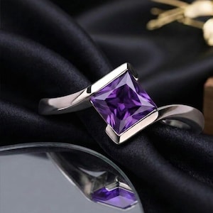May include: A silver ring with a square, deep purple gemstone. The gemstone is set in a bezel setting, and the band has a smooth, curved design. The ring is displayed on a black, textured surface.