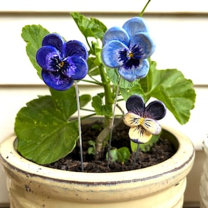 May include: A plant pot with three decorative pansy flower stakes. The pansies are blue, purple, and yellow, with green leaves. The pot is a light yellow colour.