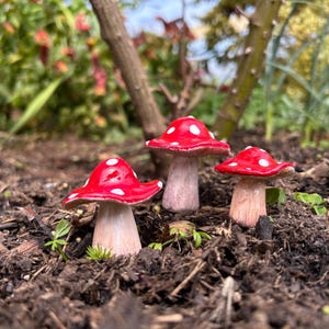 May include: Three miniature red and white spotted mushroom figurines with light pink stems are set in dark brown soil. The mushrooms are in a garden setting, with green plants and a tree trunk in the background.
