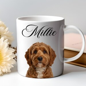 Op de afbeelding: Witte keramische mok met een portret van een bruine en witte hond en de naam "Millie" in zwart schrift. De mok heeft een gebogen handvat. De achtergrond omvat bloemen en een boek, wat een warme, uitnodigende scène creëert.