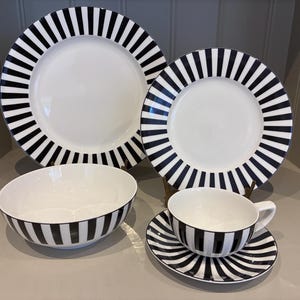 May include: A monochrome dinner service featuring white plates, a bowl, a teacup, and a saucer, all with bold black stripes. The set includes a large plate, a smaller plate. The plates, bowl, and saucer have black striped rims.
