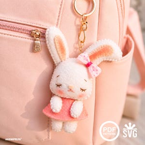 May include: A white felt bunny keyring with a pink dress and bow. The bunny has closed eyes and rosy cheeks. The keyring is attached to a gold chain and is hanging in front of a pink backpack.