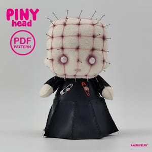 May include: A plush doll with a square, off-white head covered in grid-patterned stitching and silver pins. It features two pale eyes, a black leather-like robe, and two safety pins on the chest. Text reads PINY head, PDF PATTERN, and AKEMIFELTS.