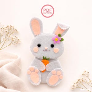 May include: A handmade felt bunny in light gray, holding an orange carrot. The bunny has pink inner ears, cheeks, and paw pads, with a purple flower on its head. The image includes the text "PDF PATTERN" in a pink circle.