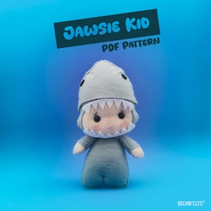 May include: A handmade felt doll wearing a gray shark costume with white teeth and black eyes. The doll has rosy cheeks and is set against a blue background. Text on the image reads "JAWSIE KID PDF PATTERN".