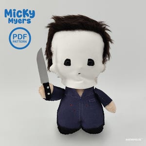 Puede incluir: Un muñeco de peluche con máscara blanca, ojos negros, cabello castaño oscuro y un mono azul marino. El muñeco sostiene un cuchillo de juguete gris en la mano derecha. El texto en la imagen dice Micky Myers, PDF PATTERN y AKEMIFELTS.