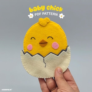 May include: A handmade felt baby chick emerging from a cracked eggshell. The chick is yellow with pink cheeks, a brown beak, and a heart-shaped headpiece. The text "baby chick PDF pattern" is above the chick.