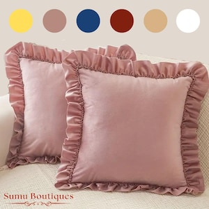 Op de afbeelding: Twee vierkante, blush-roze fluwelen sierkussens met gerimpelde randen. De kussens worden weergegeven op een lichtgekleurde bank. De tekst "Sumu Boutiques" staat onderaan de afbeelding. Het kleurenpalet omvat geel, bruin, blauw, rood, beige en wit.