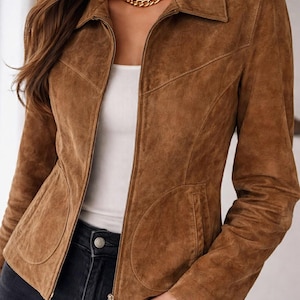 May include: A brown suede jacket with a collared neckline and a zip fastening. The jacket features two front pockets and is worn over a white top and black jeans. A gold chain necklace is visible.