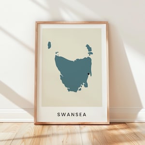 May include: A framed wall art print featuring a blue silhouette map of Tasmania on a cream background. A small black heart icon marks a specific location on the eastern coast, with the word SWANSEA printed in black capital letters at the bottom.