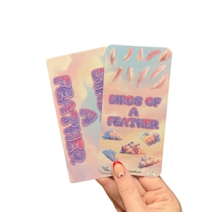 Pode incluir: Dois cartões decorativos com um design de céu pastel. Um cartão diz "FEATHER" em letras roxas em bolhas. O outro cartão diz "BIRDS OF A FEATHER" com ilustrações de penas e gráficos de nuvens. Os cartões são segurados por uma mão.