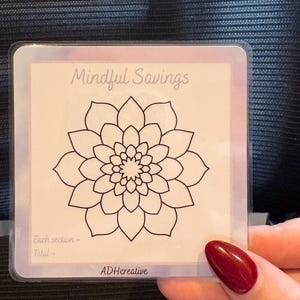 May include: A clear plastic card featuring the words "Mindful Savings" and a black floral design. The card also includes the text "Each section - Total -" and the ADHcreative logo. The card is held by a person with red nail polish.