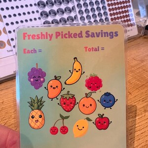 May include: A card featuring the text "Freshly Picked Savings" and cartoon illustrations of various fruits, such as a banana, pineapple, and grapes. The card has a light green background and is held by a person.