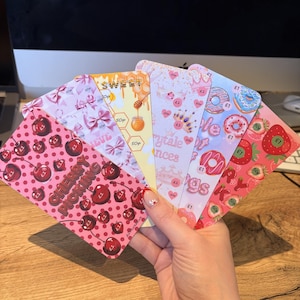 Peut inclure: Un éventail de cinq cartes rectangulaires illustrées de cerises, nœuds, miel, couronnes, beignets et fraises. Chaque carte présente des étiquettes de prix comme 50p et £1, avec des textes incluant Cherry Picking, Sweet et Crytale Princes.