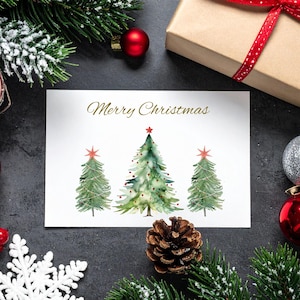 Könnte beinhalten: Eine Weihnachtskarte mit Aquarellillustrationen von drei grünen Weihnachtsbäumen, jeder mit einem roten Stern. Die Karte trägt den Text "Merry Christmas" in goldener Schrift. Die Szene umfasst festliche Dekorationen.
