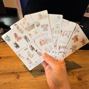 May include: A set of five white rectangular tracker cards featuring watercolor illustrations of maternity and baby-related items. The cards display icons of pregnant figures, baby clothes, furniture, and accessories with text including Maternity Clothes Fund.