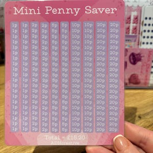 May include: A pink Mini Penny Saver card with a grid for saving coins. The card has the text "Mini Penny Saver" at the top and a grid of boxes with coin values. The total is £15.20.