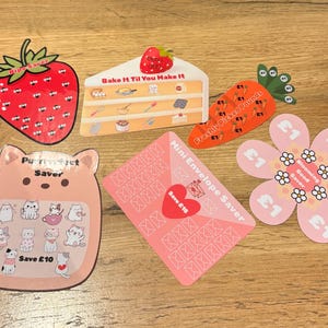 May include: A selection of themed money-saving trackers. Includes a strawberry, cake slice, carrot, cat-shaped tracker, flower, and pink envelope. Each tracker has spaces for saving money, with text like "Save £10" and "Mini Envelope Saver."