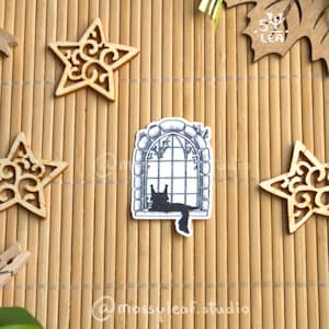 Puede incluir: Una colección de estrellas decorativas de madera, una etiqueta en forma de hoja y una pegatina de un gato en una ventana están dispuestas sobre una esterilla de bambú. La etiqueta de la hoja está atada con cinta verde. También hay pequeñas pinzas de madera.