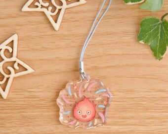 May include: A clear acrylic charm featuring a small, round, orange-red flame character with a smiling face, surrounded by light blue and pink decorative accents. The charm is attached to a thin, light blue cord loop for hanging purposes.