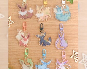 May include: A collection of nine clear acrylic keychains featuring various stylized creatures with plant-like elements, each attached to a colored plastic lobster clasp. The designs include brown, yellow, blue, pink, dark gray, and purple characters.