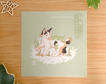 May include: A square art print on a light green background featuring a calico Japanese bobtail cat walking through grass. The text reads Japanese bobtail cat and Japanese characters. A small logo with the text LEA is visible in the bottom left corner.