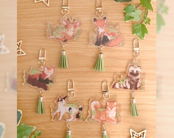 May include: Six clear acrylic keychains featuring illustrated animals including a deer, fox, raccoon, cat, and dog, each adorned with green foliage accents. Each keychain includes a gold-toned lobster clasp and a small sage green faux leather tassel.