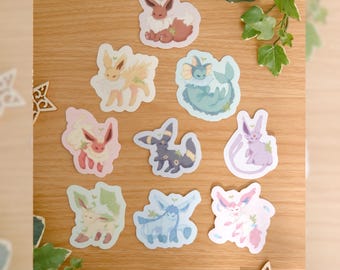 May include: A collection of nine die-cut vinyl stickers featuring various stylized creatures with plant-like features, including brown, yellow, blue, pink, and purple color palettes, arranged on a wooden surface with the text by mossyleaf studio.
