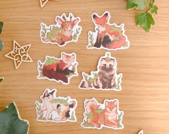 May include: A collection of six die-cut stickers featuring illustrated animals including a deer, foxes, and a cat, each adorned with green foliage. The stickers are arranged on a wooden surface alongside three wooden star cutouts and green leaves.