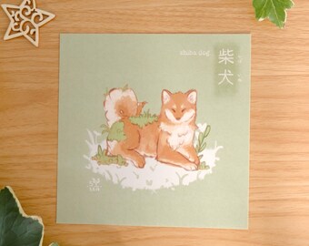 May include: A square art print on light green paper featuring a brown and white Shiba Inu dog lying down in grass. The text "shiba dog" and Japanese characters are printed in the upper right corner, with a small logo in the lower left corner.