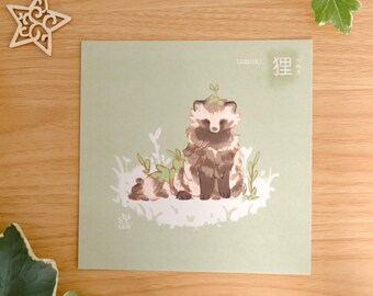 May include: A square art print on a light green background featuring a brown and white tanuki illustration. The tanuki sits among small green plants with a sprout on its head. The text tanuki and the Japanese kanji 狸 are printed in the top right corner.