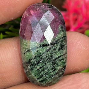 Könnte beinhalten: Ein ovaler Edelstein mit facettierter Oberseite. Der Edelstein weist eine Mischung aus Farben auf, darunter tiefes Rosa, Grün und Schwarz. Die Oberseite des Edelsteins hat ein reflektierendes, geometrisches Muster. Der Stein wird zwischen Fingern gehalten.