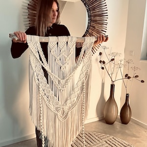 Könnte beinhalten: Cremefarbener Makramee-Wandbehang mit aufwendigen geometrischen Mustern und langem Fransen. Die Textilkunst ist an einem natürlichen Holzdübel aufgehängt. Ein dekoratives Stück für die Boho- oder moderne Inneneinrichtung.