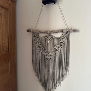 Può includere: Un arazzo da parete in macramè grigio con un'asta di legno e frange. Il macramè presenta un design geometrico dettagliato nella parte superiore e un accento di perline. L'arazzo è sospeso da un apparecchio di illuminazione cilindrico nero.