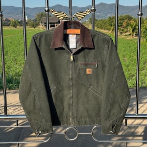 Puede incluir: Chaqueta de trabajo verde oscuro con cuello de pana marrón y cierre de cremallera. La chaqueta tiene un pequeño bolsillo cuadrado en el pecho y un parche con el logo de Carhartt. La chaqueta está colgada en una percha decorativa.