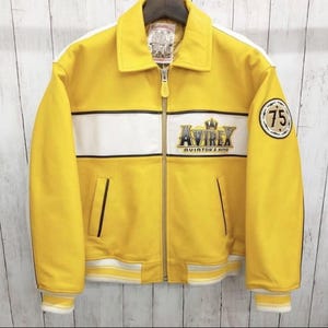 May include: Yellow leather jacket with white stripes and accents. Features the "AVIREX AVIATOR'S KING" logo and a "75" patch. Includes a zipper closure and a collar. The jacket has a classic bomber style.