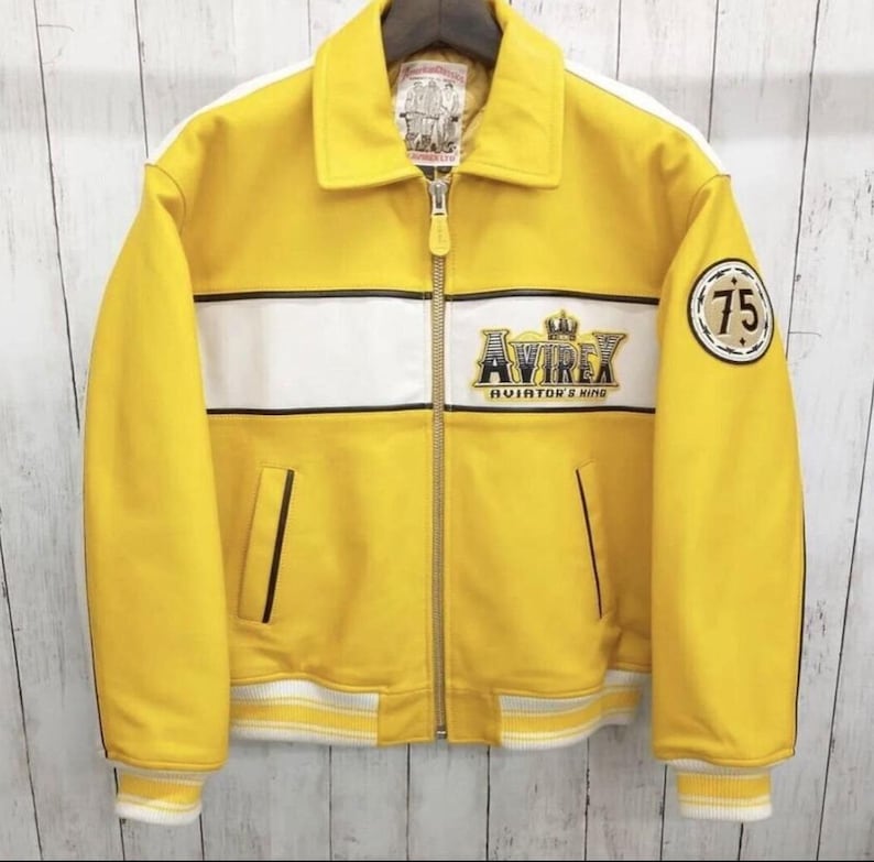 May include: Yellow leather jacket with white stripes and accents. Features the "AVIREX AVIATOR'S KING" logo and a "75" patch. Includes a zipper closure and a collar. The jacket has a classic bomber style.