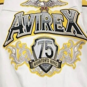 May include: Close-up of a white jacket with embroidered details. The jacket features the word "AVIREX" in a bold, black and yellow design. Below, a shield displays "75" and "AVIATOR'S KING" in a banner, with an eagle emblem at the top.