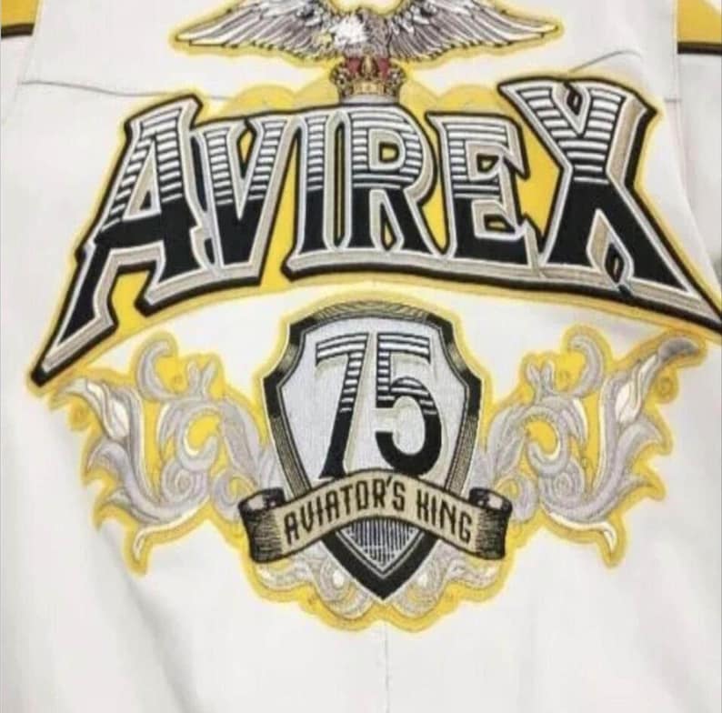 May include: Close-up of a white jacket with embroidered details. The jacket features the word "AVIREX" in a bold, black and yellow design. Below, a shield displays "75" and "AVIATOR'S KING" in a banner, with an eagle emblem at the top.
