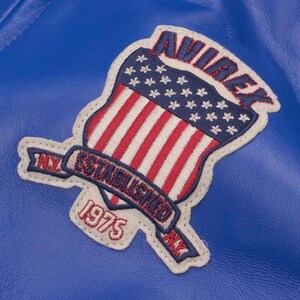 May include: Close-up of a blue leather jacket with a stitched patch. The patch features a shield design with the American flag, the word "AVIREX" in red, and the text "ESTABLISHED 1975 NY".