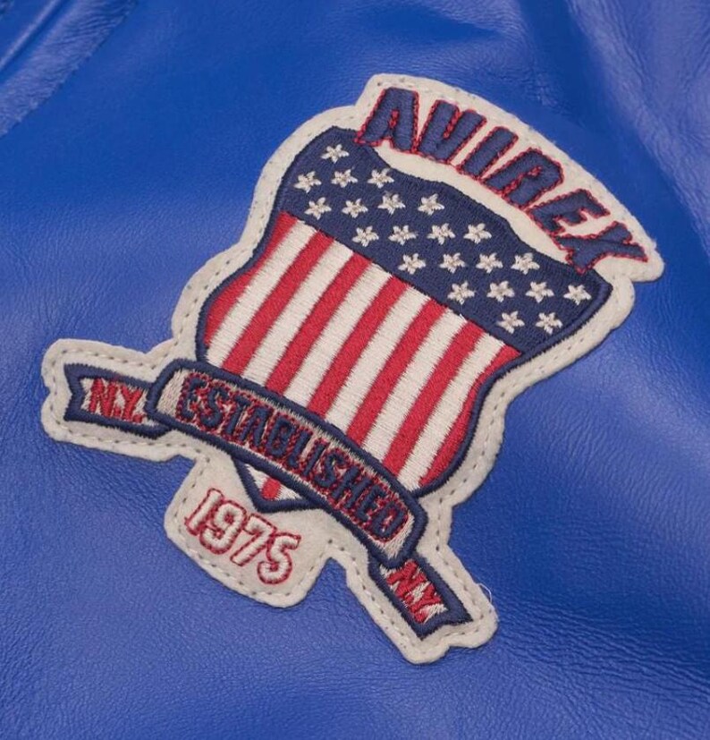 May include: Close-up of a blue leather jacket with a stitched patch. The patch features a shield design with the American flag, the word "AVIREX" in red, and the text "ESTABLISHED 1975 NY".