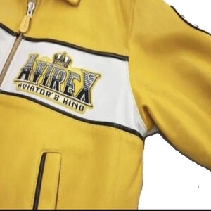 May include: Yellow jacket with white and black accents. The jacket features a zipper closure and a patch on the chest with the text "AVIREX AVIATOR'S KING" in a stylized font. The jacket has a classic bomber style.