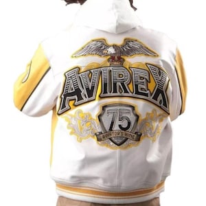 May include: White and yellow varsity jacket with the word "AVIREX" in large, black, outlined letters on the back. An eagle graphic is above the text, and a shield with the number "75" is below. The jacket has a hood and yellow accents.