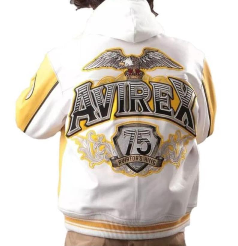May include: White and yellow varsity jacket with the word "AVIREX" in large, black, outlined letters on the back. An eagle graphic is above the text, and a shield with the number "75" is below. The jacket has a hood and yellow accents.