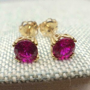 Puede incluir: Un par de pendientes de oro con gemas de color magenta vibrante. Las gemas de corte redondo están engastadas en una montura de cuatro garras. Los pendientes se muestran sobre una superficie clara y texturizada.