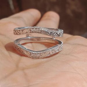 May include: A silver ring guard with an ornate design. The ring features a curved shape with intricate detailing and small, sparkling stones. The design includes delicate patterns and a polished finish, creating an elegant look.